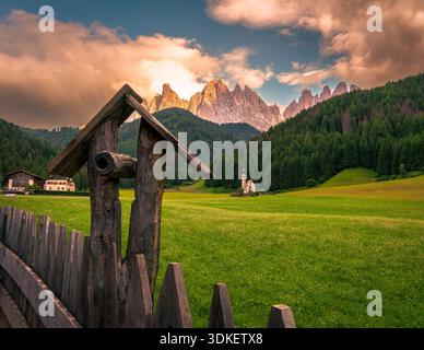Im Vordergrund steht ein rustikales Vogelhaus aus Holz mit Blick auf eine lebendige grüne Wiese mit Wildblumen. In der Mitte des Erdgeschosses befindet sich eine bezaubernde Kirche Stockfoto