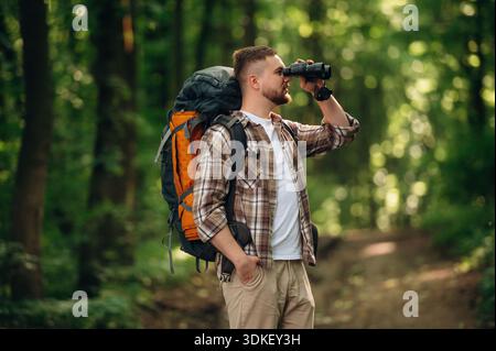Seitenansicht, stehend, Fernglas halten. Männlicher Tourist ist mit Rucksack im Wald. Stockfoto
