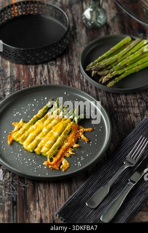 Spargelspeere bedeckt mit geschmolzenem Käse auf einem dunklen Teller, verzehrfertig Stockfoto
