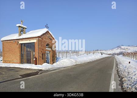 Kleine rote Kapelle am Straßenrand zwischen Weinbergen, die von weißem Schnee bedeckt sind, unter klarem blauem Winterhimmel im Piemont, Norditalien Stockfoto