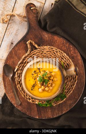 Ein warmer, von oben nach unten leuchtender gelber Gemüsepüree in einem Keramiktopf. Die rustikale Szene zeigt natürliche Texturen aus einem Holzbrett und gewebtem Tischset Stockfoto