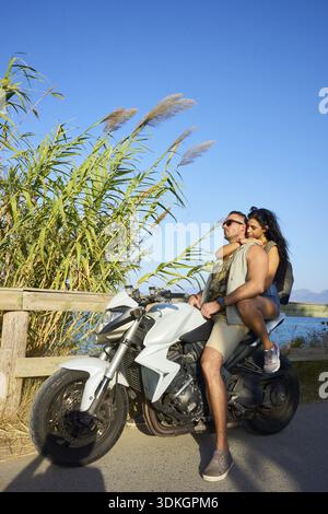 Entspannter Ausflug mit einem Paar, das an einem sonnigen Tag neben hohen Gräsern auf einem Motorrad fährt, das Abenteuer mitnimmt, Paar auf einem Motorrad auf einem landschaftlich reizvollen Ro Stockfoto