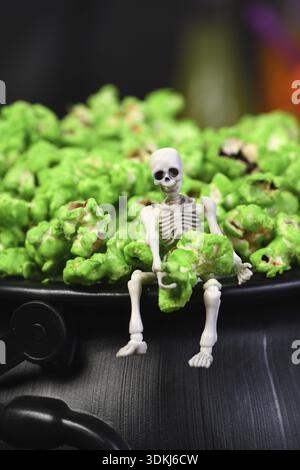 Essen, Ein Skelett sitzt am Rand eines Kessels und hält in den Händen ein grünes süßes Popcorn. Nahaufnahme. Halloween-Leckereien Stockfoto