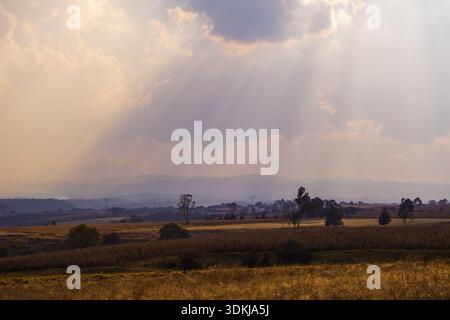 Sonnenstrahlen durchbrechen Wolken über ländliche Landschaft mit Feldern und fernen Bergen, dramatischem Himmel und natürlichem Licht Stockfoto