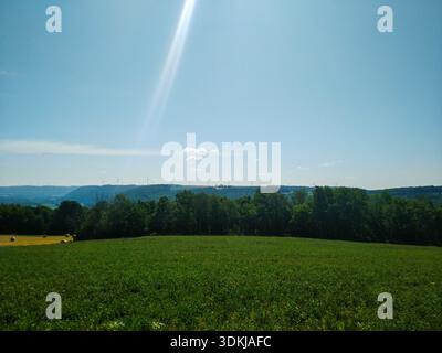 Sonnenstrahl bricht durch klaren Himmel über grünen Feldern mit Bäumen und fernen Hügeln. Stockfoto