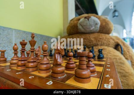 Nahaufnahme eines hölzernen Schachbretts mit geschnitzten Schachfiguren an einer Straßenecke, mit einem großen Teddybären im Hintergrund. Stockfoto
