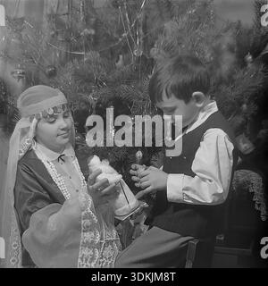 Slawjansk, Ukraine, 1970er Jahre Ein authentisches Archivfoto von Geschwistern, die Neujahr feiern. Kravelis Inessa Michailowna, gekleidet in einem handgefertigten Schneemädchen Kostüm, zeigt ihrem Bruder Kravelis Dmitri Michailowitsch eine Spielzeugfigur Ded Moroz. Der Junge sitzt auf einem Stuhl, trägt eine Weste und ein weißes Hemd. Sie posieren neben einem Weihnachtsbaum, der mit Vintage-Glasornamenten und Lametta verziert ist. Dieser hochauflösende Scan erfasst die festliche Atmosphäre, die familiäre Bindung und die Traditionen im friedlichen Donbass während der Ära des Lebens vor dem Krieg. Stockfoto