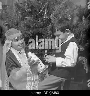 Slawjansk, Ukraine, 1970er Jahre Ein authentisches Archivfoto von Geschwistern, die Neujahr feiern. Kravelis Inessa Michailowna, gekleidet in einem handgefertigten Schneemädchen Kostüm, zeigt ihrem Bruder Kravelis Dmitri Michailowitsch eine Spielzeugfigur Ded Moroz. Der Junge sitzt auf einem Stuhl, trägt eine Weste und ein weißes Hemd. Sie posieren neben einem Weihnachtsbaum, der mit Vintage-Glasornamenten und Lametta verziert ist. Dieser hochauflösende Scan erfasst die festliche Atmosphäre, die familiäre Bindung und die Traditionen im friedlichen Donbass während der Ära des Lebens vor dem Krieg. Stockfoto