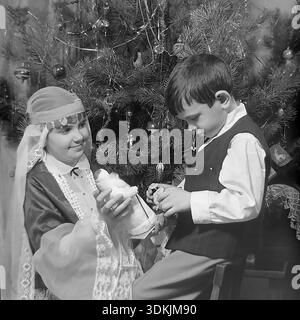 Slawjansk, Ukraine, 1970er Jahre Ein authentisches Archivfoto von Geschwistern, die Neujahr feiern. Kravelis Inessa Michailowna, gekleidet in einem handgefertigten Schneemädchen Kostüm, zeigt ihrem Bruder Kravelis Dmitri Michailowitsch eine Spielzeugfigur Ded Moroz. Der Junge sitzt auf einem Stuhl, trägt eine Weste und ein weißes Hemd. Sie posieren neben einem Weihnachtsbaum, der mit Vintage-Glasornamenten und Lametta verziert ist. Dieser hochauflösende Scan erfasst die festliche Atmosphäre, die familiäre Bindung und die Traditionen im friedlichen Donbass während der Ära des Lebens vor dem Krieg. Stockfoto