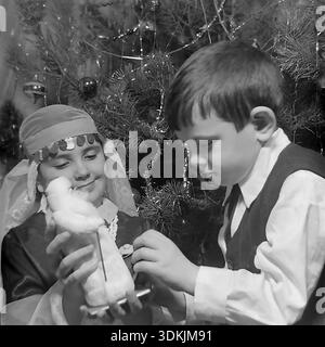 Slawjansk, Ukraine, 1970er Jahre Ein authentisches Archivfoto der Geschwister Kravelis Inessa Michailowna und Kravelis Dmitri Michailowitsch, die Neujahr feiern. Das Mädchen trägt ein handgefertigtes Schneemädchen-Kostüm mit Schleier und hält eine Ded Moroz-Figur. Ihr Bruder, der eine Weste und ein weißes Hemd trägt, sieht das Spielzeug neugierig an. Sie stehen neben einem Weihnachtsbaum, der mit Vintage-Glasschmuck verziert ist. Dieser hochauflösende Scan erfasst die festliche Atmosphäre, die familiäre Bindung und die Traditionen im friedlichen Donbass während der Ära des Lebens vor dem Krieg. Stockfoto