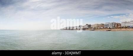 Ein breiter Blick auf die Küstenstadt mit ruhigem Meer, Sandstrand und einer belebten Uferpromenade mit Gebäuden und einem Riesenrad am Horizont. Hell, sonnig, Stockfoto