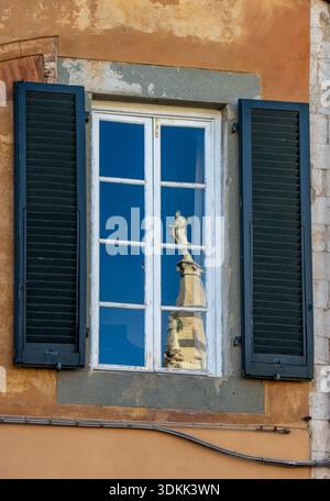 Detalle de una ventana tradicional en Pisa, Italia, contraventanas verdes y fachada de tonos cálidos, reflejando una estatua histórica en el Cristal. Stockfoto