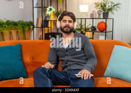 Ein indischer Mann zu Hause trägt kabellose Kopfhörer spielt Naturgeräusche, Atmen, Meditieren und Lächeln. Ein Araber auf dem Sofa hört ruhigen Lärm zu, zeigt entspannte Stimmung und achtsame Pause Stockfoto