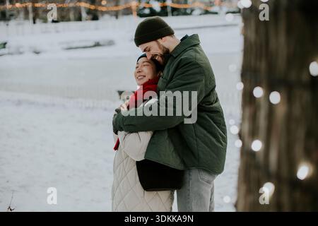 Eine Koreanerin und ein Kaukasier lächeln sich in einem verschneiten Park herzlich an. Sie stehen dicht beieinander, umgeben von Lichtern und Winterlandschaften Stockfoto