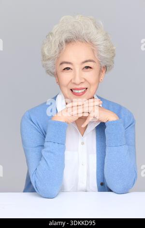 Fröhliche ältere asiatische Frau mit kurzen silbernen Haaren in blauer Strickjacke, die Glück und gesunden Lebensstil zum Ausdruck bringt, Studio-Porträt auf einem weißen bac Stockfoto