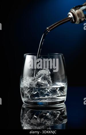 Kristallklarer Wodka oder Gin wird über Eis in ein Glas gegossen. Wodka mit Eis auf schwarzem reflektierendem Hintergrund. Stockfoto