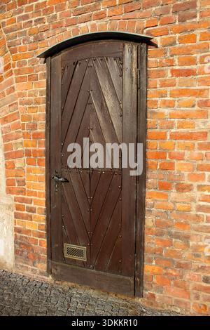Alte Holztür mit Metallscharnieren in der historischen roten Backsteinwand mit Textur von Mauerwerk und veralteten architektonischen Details. Stockfoto