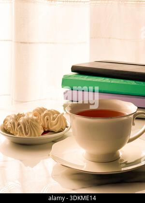 Tee mit Zephyr Marshmallows. Gemütliche Teezeit mit einer Tasse heißem schwarzem Tee und weichen, luftigen russischen zephyr Marshmallows auf einem Leuchttisch in der Nähe des Fensters. Stockfoto