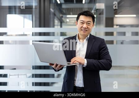 Asiatischer Geschäftsmann im Anzug lächelt selbstbewusst, während er auf einem silbernen Laptop in einem hellen, modernen Büroflur mit mattierten Glasabtrennungen und klaren architektonischen Linien tippt Stockfoto