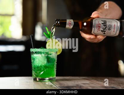 Ein Mann gießt ein Getränk aus einer Flasche in ein Glas. Das Getränk ist grün und hat einen Limettenkeil Stockfoto