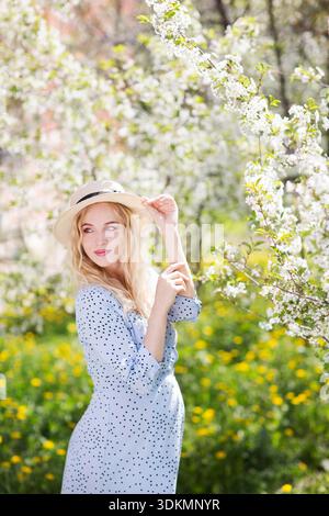 Eine schöne junge schwangere Frau in einem Kleid mit Strohhut geht durch den Frühlingsgarten. Glückliche Schwangerschaft. || Modell freigegeben Stockfoto