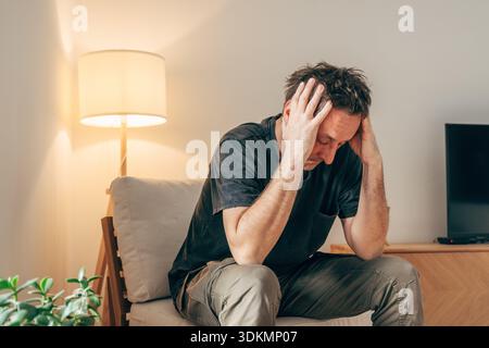 Mann in emotionaler Not sitzend auf einem Stuhl, Kopf in Händen, warme Innenbeleuchtung, selektiver Fokus Stockfoto