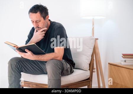 Authentischer reifer Erwachsener, der sich tief konzentriert, während er ein offenes Buch liest, Hand am Kinn. Wissen, Weisheit, Reflexion, Bildung, Literaturthemen. Wählen Sie Aus Stockfoto
