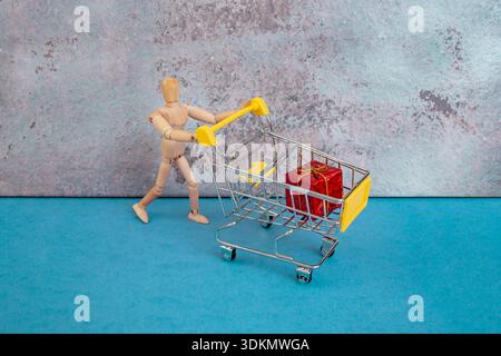 Holzpuppe schiebt einen Einkaufswagen mit einem roten Geschenk, Valentinstag-Geschenk-Konzept Stockfoto