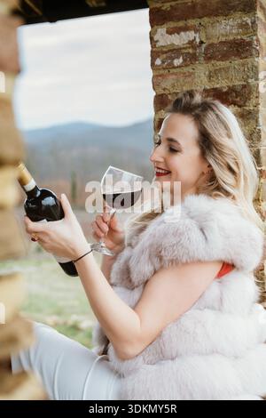 Frau in einer Pelzweste genießt draußen ein Glas Rotwein, das Entspannung und Vergnügen zeigt. Sie lächelt und schätzt den Moment vor einem natürlichen Rücken Stockfoto