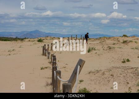 Silhouette eines einsamen Wanderers, der durch Sanddünen mit einem hölzernen Seilzaun im Vordergrund und majestätischen Bergen am Horizont spaziert Stockfoto