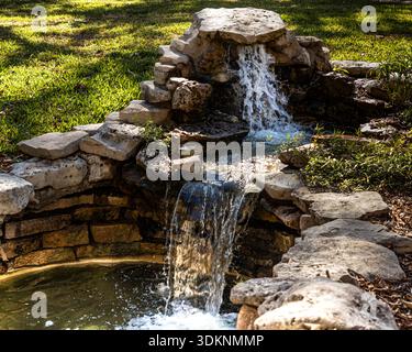 Kleiner Steingarten Wasserfall fließt in einen ruhigen Hinterhofteich mit Sonnenlicht, das auf dem Wasser reflektiert Stockfoto