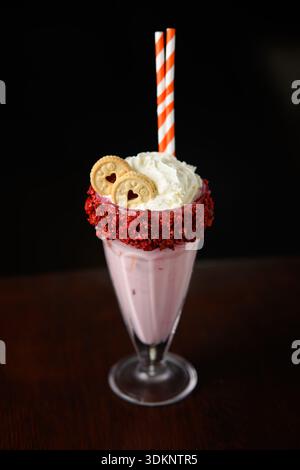 Ein rosafarbener Milchshake mit einem herzförmigen Keks drauf. Der Milchshake wird in einem Glas mit rotem und weißem Strohhalm serviert Stockfoto