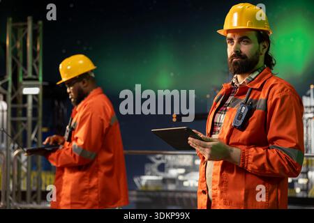 Umweltbeauftragter auf Offshore-Plattform, der die CO2-Bilanz mithilfe eines Tablets unter Nordlichtern verfolgt. Arbeiter am Bohrgerät sorgen für nachhaltige Bohrpraktiken während aurora borealis Stockfoto