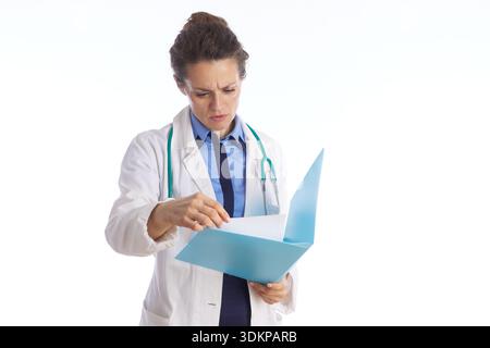 Eine fokussierte Ärztin in einem weißen Labormantel und Stethoskop prüft Patientendokumente in einem hellblauen Ordner vor weißem Studiohintergrund. Stockfoto