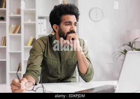 Der müde Indianer nimmt die Brille ab, leidet unter schmerzhaften Empfindungen, hat Probleme mit Astigmatismus. Ein Mann, der nach einem langen Arbeitstag auf der Komm gestresst hat Stockfoto