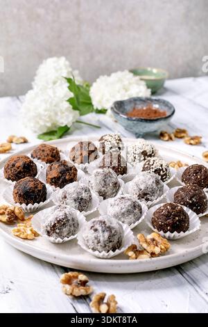 Sortiment hausgemachte Schokolade rohe vegane Kugeln Trüffel mit verschiedenen Belägen auf weißem Teller. Kakaopulver, Walnüsse verstreut. Kleine Schüsseln mit Stockfoto
