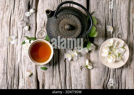 Gusseiserne Teekanne mit Tasse heißem Tee Jasmin Blumen auf rustikalem hölzernem Hintergrund. Traditionelle asiatische Teezeremonie mit natürlicher Blumendekoration. Stockfoto