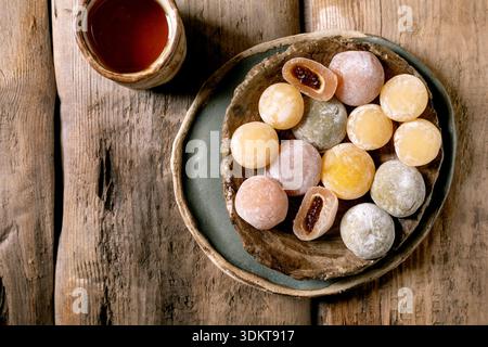 Bunte Mochi-Kugeln mit süßer Füllung auf Keramikplatte neben Teetasse auf rustikalem Holztisch. Traditionelles japanisches Süßwarendessert. Stockfoto