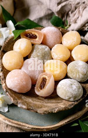 Farbenfrohe traditionelle japanische Mochi-Reiskuchen mit süßer Füllung auf rustikalem Holzteller mit weißen Blumen und grünen Blättern Stockfoto