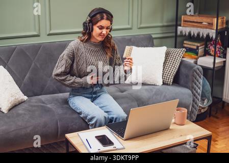 Die junge Frau sitzt auf dem Bett im Wohnzimmer und hat einen Online-Arzttermin mit Laptop. Junge Frau, die Medikamente nimmt, während sie an Lapto arbeitet Stockfoto
