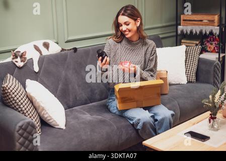 Glückliche Frau, die sich Parfüms ansieht, die sie online gekauft hat, und bekam ein Paket geliefert. Ihre Katze liegt auf dem Sofa neben ihr. Stockfoto
