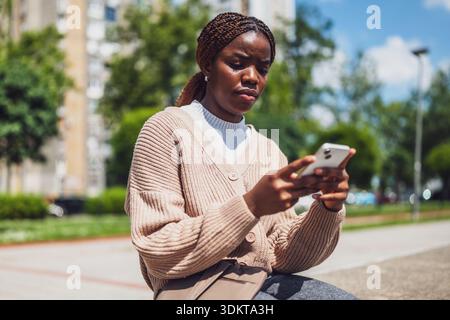 Die mürrische afroamerikanische Frau ist besorgt, wenn sie ihr Smartphone für Online-Nachrichten benutzt. Stockfoto