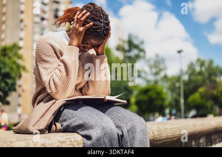 Ängstliche afroamerikanische Frau sitzt im Stadtpark, tief in Gedanken und schreibt ihr Tagebuch. Stockfoto