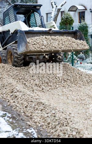 Ein kleines Baufahrzeug wirft Kies auf schneebedeckten Boden in der Nähe eines Hauses mit Bogenfenstern, Topfpflanzen und einem Zaun in winterlicher Umgebung ab. Stockfoto