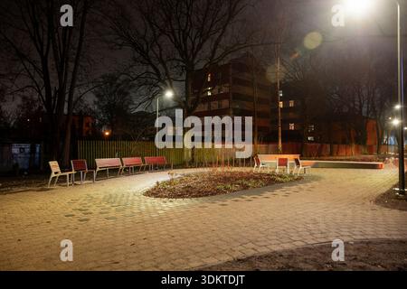 Ein kleiner Stadtpark bei Nacht mit einem geschwungenen Weg, roten und weißen Bänken, karger Vegetation und mehrstöckigen Gebäuden im Hintergrund. Stockfoto