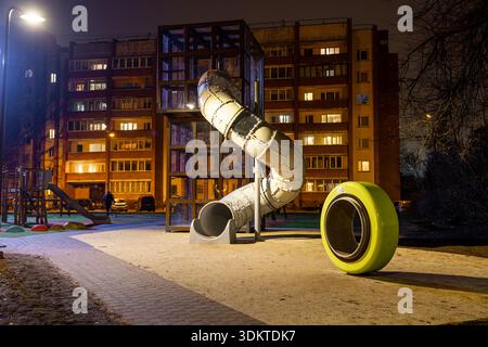 Nächtlicher Blick auf einen Spielplatz mit einer hohen Spiralrutsche, gelbem Spielelement, gepflastertem Gang und einem beleuchteten Wohngebäude im Hintergrund. Stockfoto