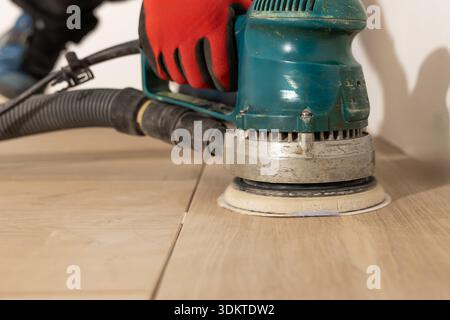 Eine Person verwendet eine blaue elektrische Bodenschleifmaschine mit einem schwarzen Vakuumschlauch, um Holzböden zu polieren. Rote Handschuhe und Steppnähte betonen Präzision. Stockfoto