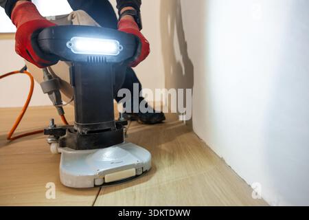 Nahaufnahme eines Arbeiters mit einer Bodenpoliermaschine auf einem hellen Holzboden im Innenbereich, mit roten Handschuhen, einem orangefarbenen Netzkabel und einer sichtbaren weißen Wand. Stockfoto