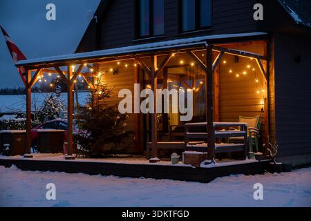 Eine hölzerne Hausterrasse mit LED-Lichtern, einem Weihnachtsbaum, Gartenmöbeln und schneebedeckter Umgebung in ländlicher Umgebung. Stockfoto
