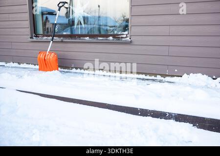 Eine orangene Schneeschaufel liegt an der Wand eines Gebäudes mit horizontaler Abschirmung. Ein zerrissenes Fenster spiegelt nahe gelegene Strukturen wider. Schnee bedeckt den Boden mit Stockfoto